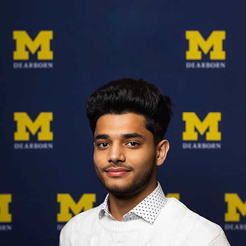 Person smiling at the camera with short hair and a white shirt on with a UM-Dearborn logo background.