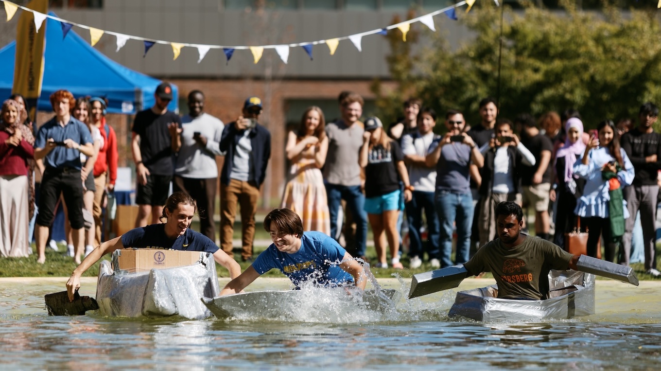 Three participants race homemade cardboard boats across a body of water in front of a cheering crowd during a campus homecoming event.