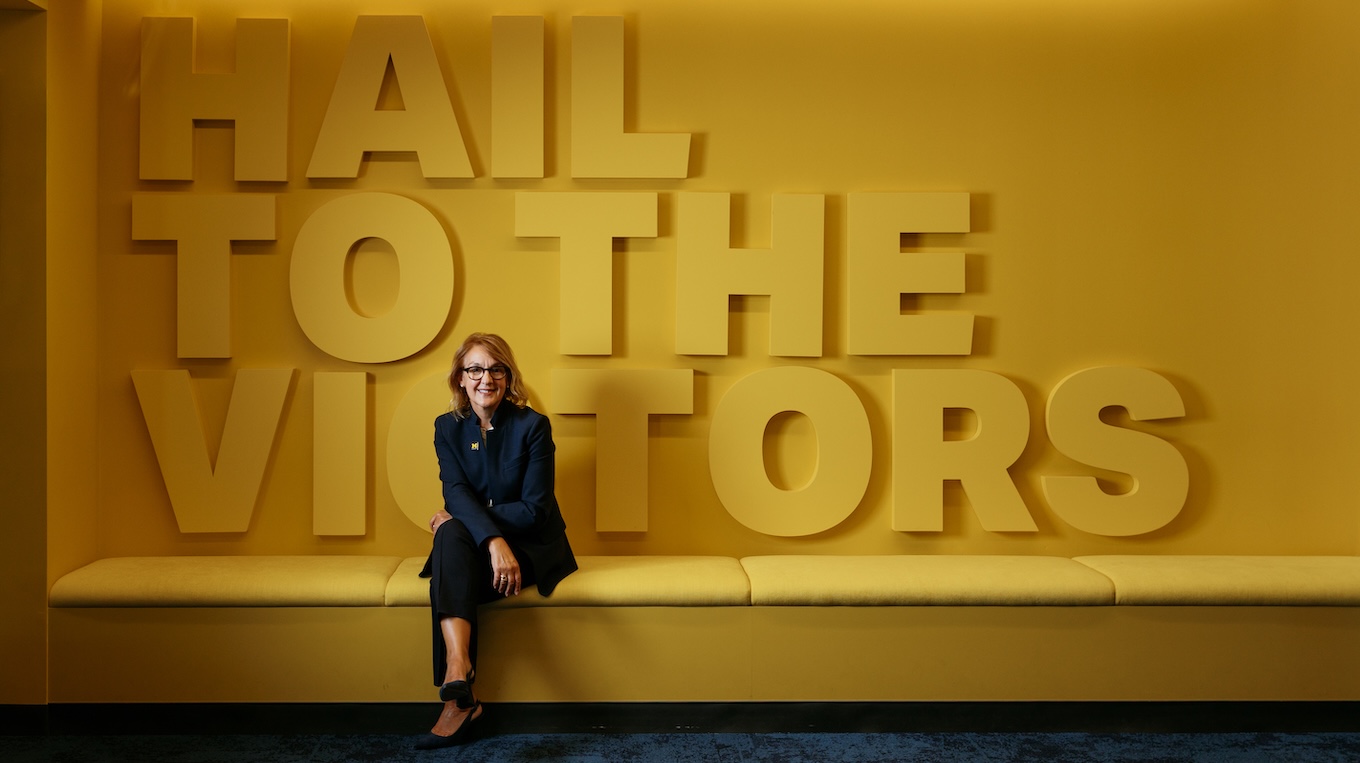 Chancellor Gabriella Scarlatta sits on a yellow bench in front of a wall featuring large 3D block letters that spell out "HAIL TO THE VICTORS."