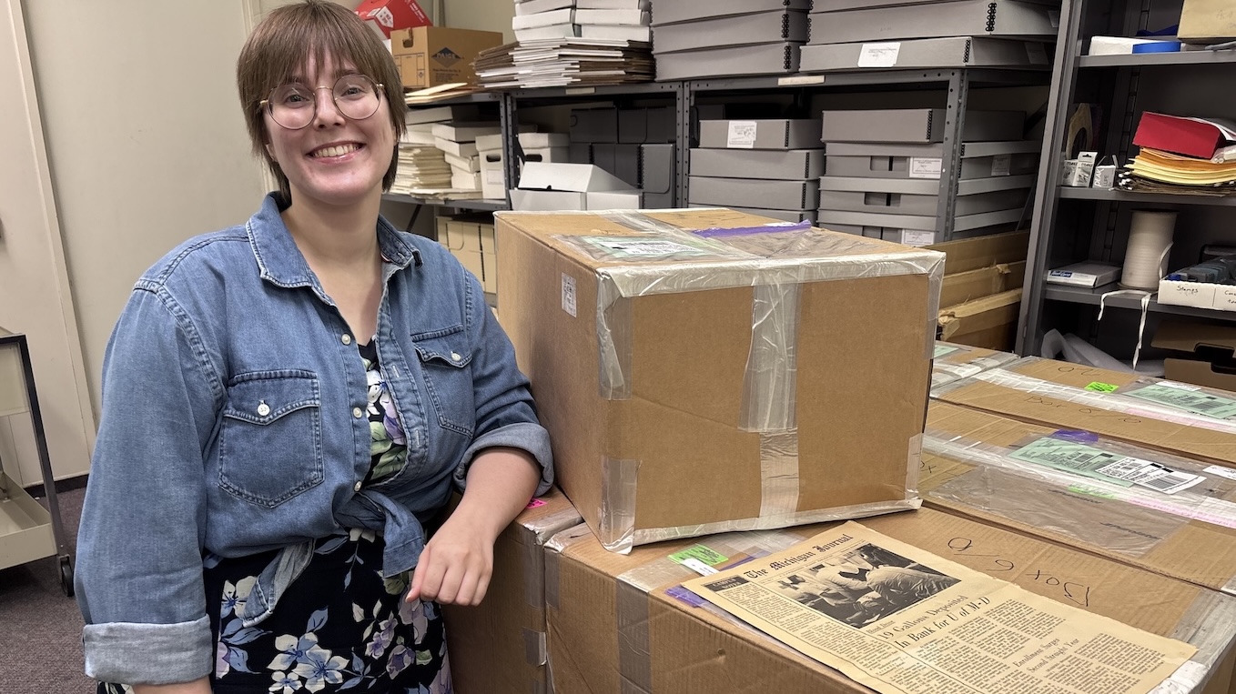 A smiling person with short brown hair, glasses, and a denim shirt over a floral dress standing next to large cardboard boxes in an archive or storage room filled with shelves of document boxes