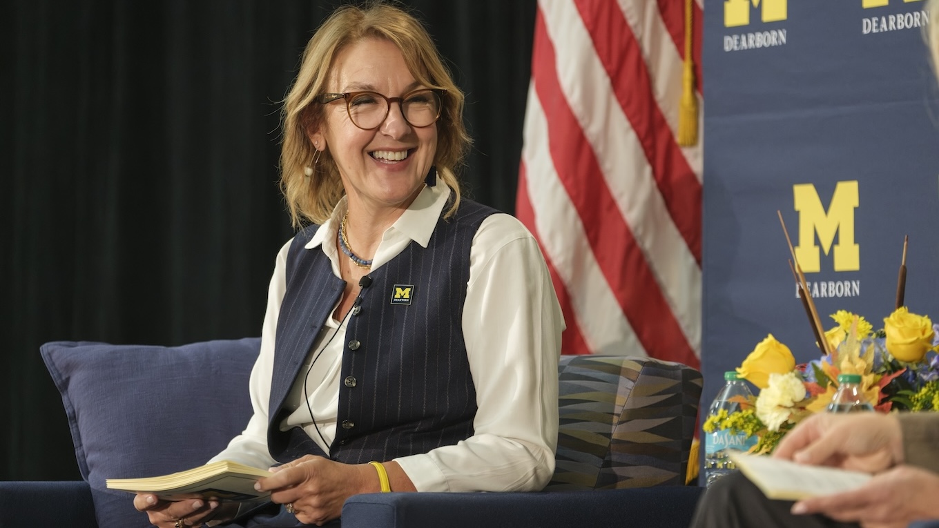 A woman wearing glasses, a white blouse and a pinstriped navy vest smiles warmly while sitting on a armchair holding a small booklet during an event with a University of Michigan-Dearborn backdrop and an American flag.