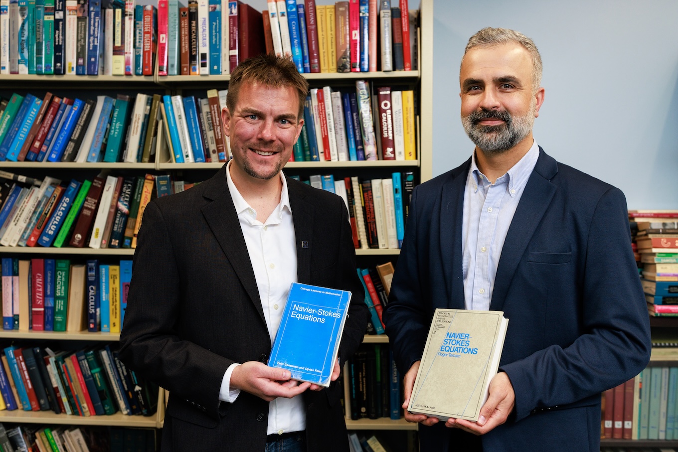 Two men standing in an office in front of bookshelves filled with math textbooks, each holding a book titled "Navier-Stokes Equations."