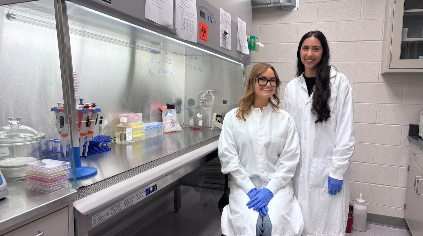Two smiling women wearing white lab coats stand together in a laboratory next to a biosafety cabinet stocked with lab equipment.   