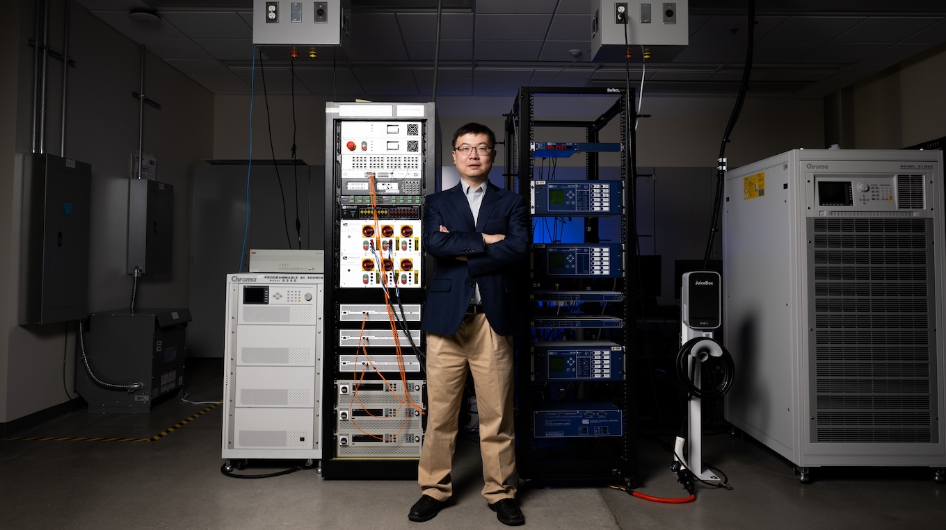 A man wearing glasses, a dark blazer, and tan trousers stands with his arms crossed between tall electronic equipment racks in a dimly lit laboratory or engineering facility. He is surrounded by scientific hardware, including programmable power supply units labeled "Chroma" and an EV charging station.   
