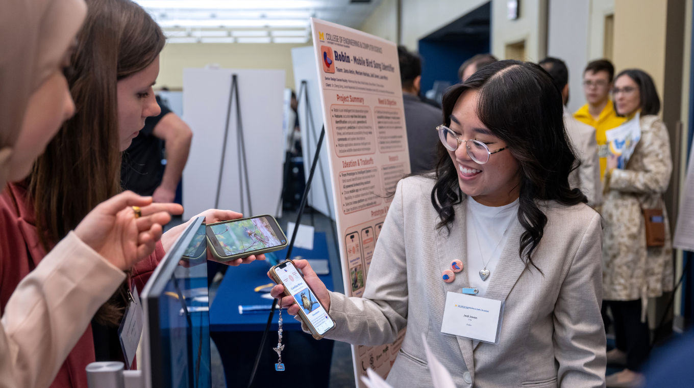 A smiling student demonstrates a mobile app on her phone to two attendees during a crowded University of Michigan-Dearborn research poster session.