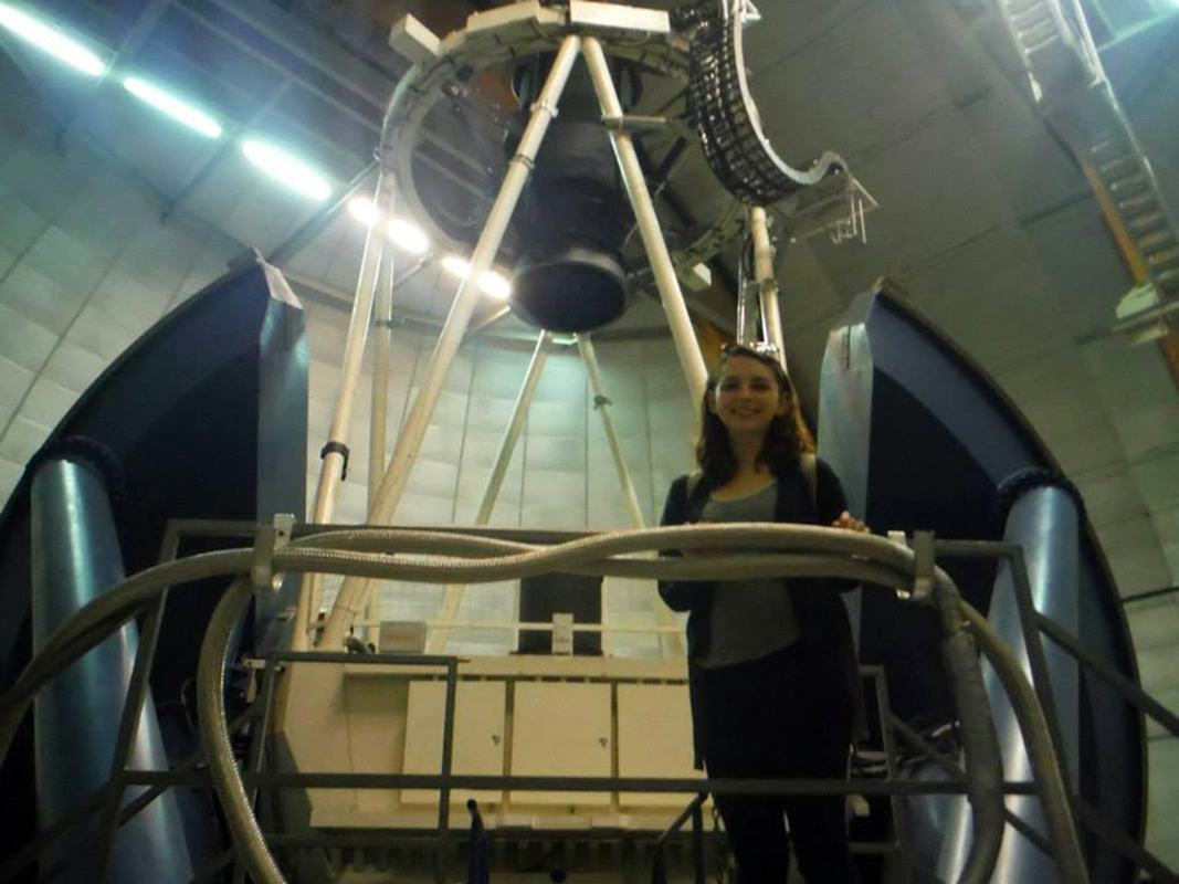 Brittany Howard is a young white woman with shoulder length brown hair. She is standing in an observatory and is wearing a gray tank underneath a black cardigan. Her glasses are set over her head.
