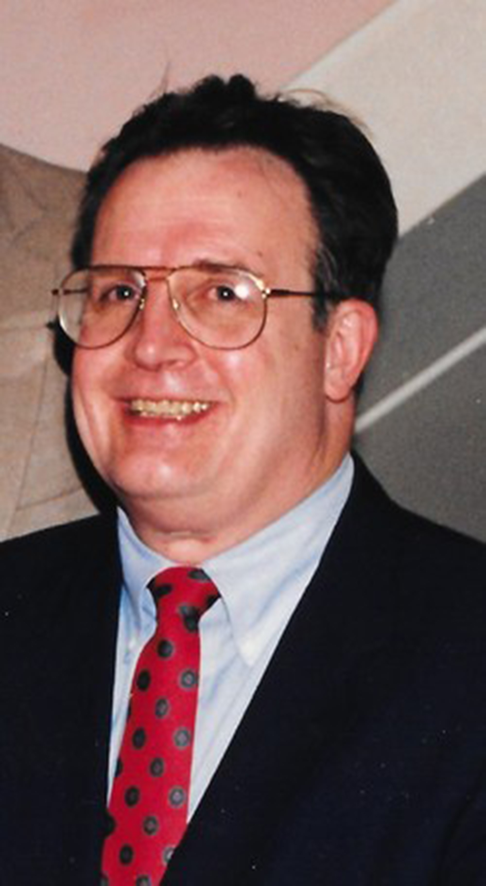 The late Donald Rossin was an elderly white man with short, jet black hair and a pair of aviator glasses. He is smiling in this photo, wearing a navy suit jacket over a white button up and a red/black polka dot tie.