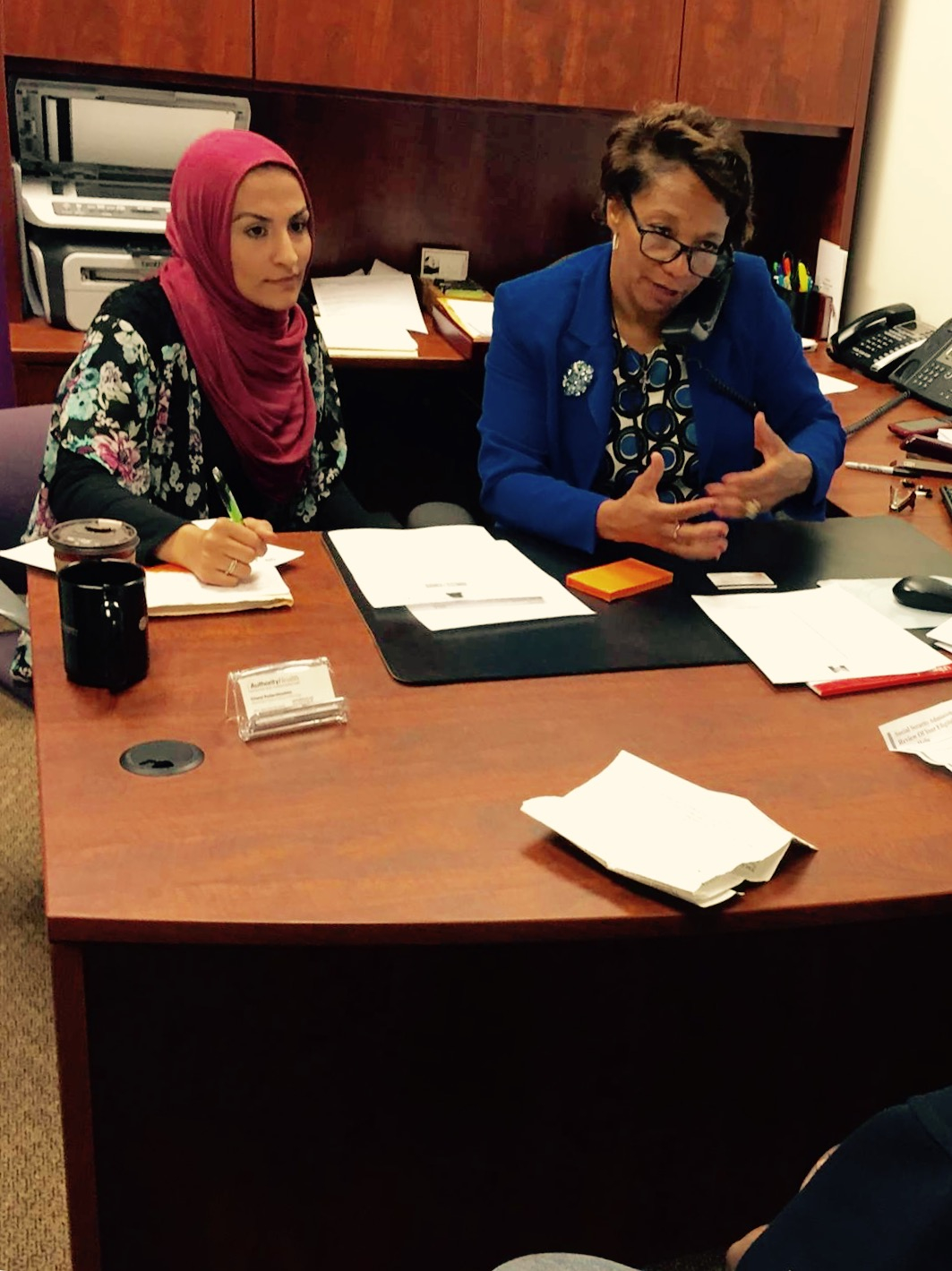 Gadah Sharif and Julie Roddy in an office sitting behind a desk. Julie is on the phone while Gadah is writing on a notepad.