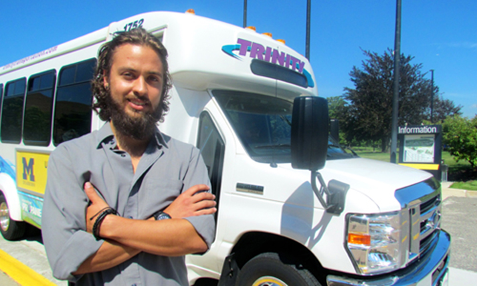 Thomaz is a young, Latinx man with tanned skin, green eyes, and dark brown, chin-length curly hair & facial hair. He is smiling, standing in front of a UM-Dearborn Trinity Bus with his arms crossed. Thomaz is wearing a light-gray button down with the sleeves rolled up.