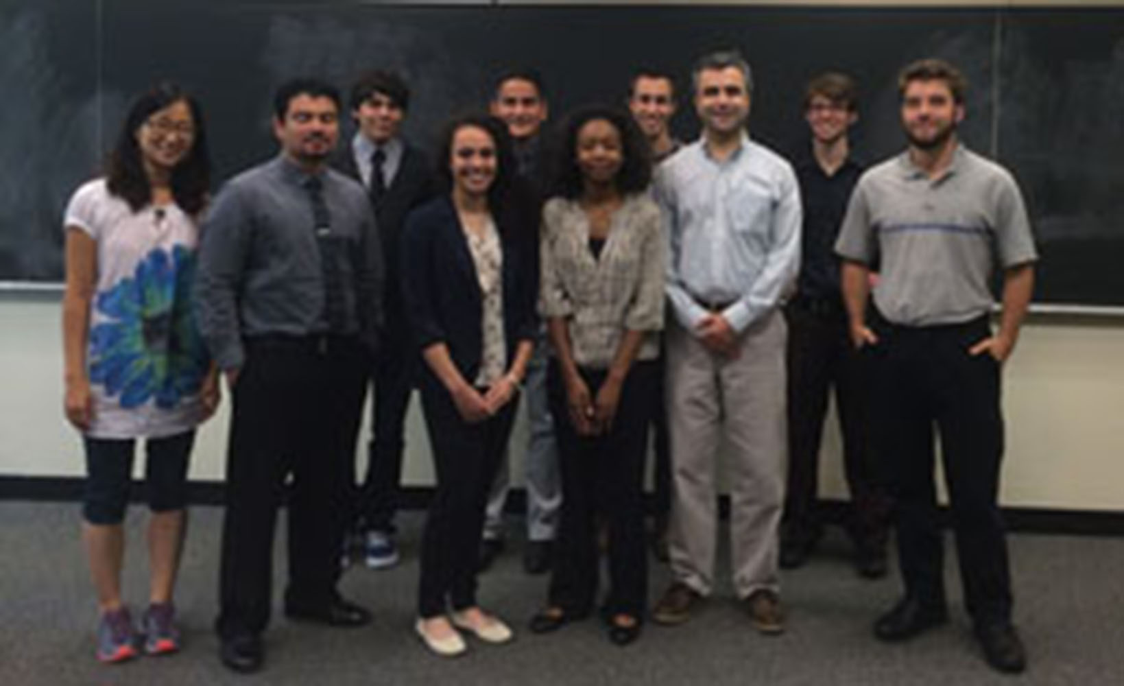 A group of students from the mathematics summer research program. They are standing in the front of a classroom with a black board behind them.