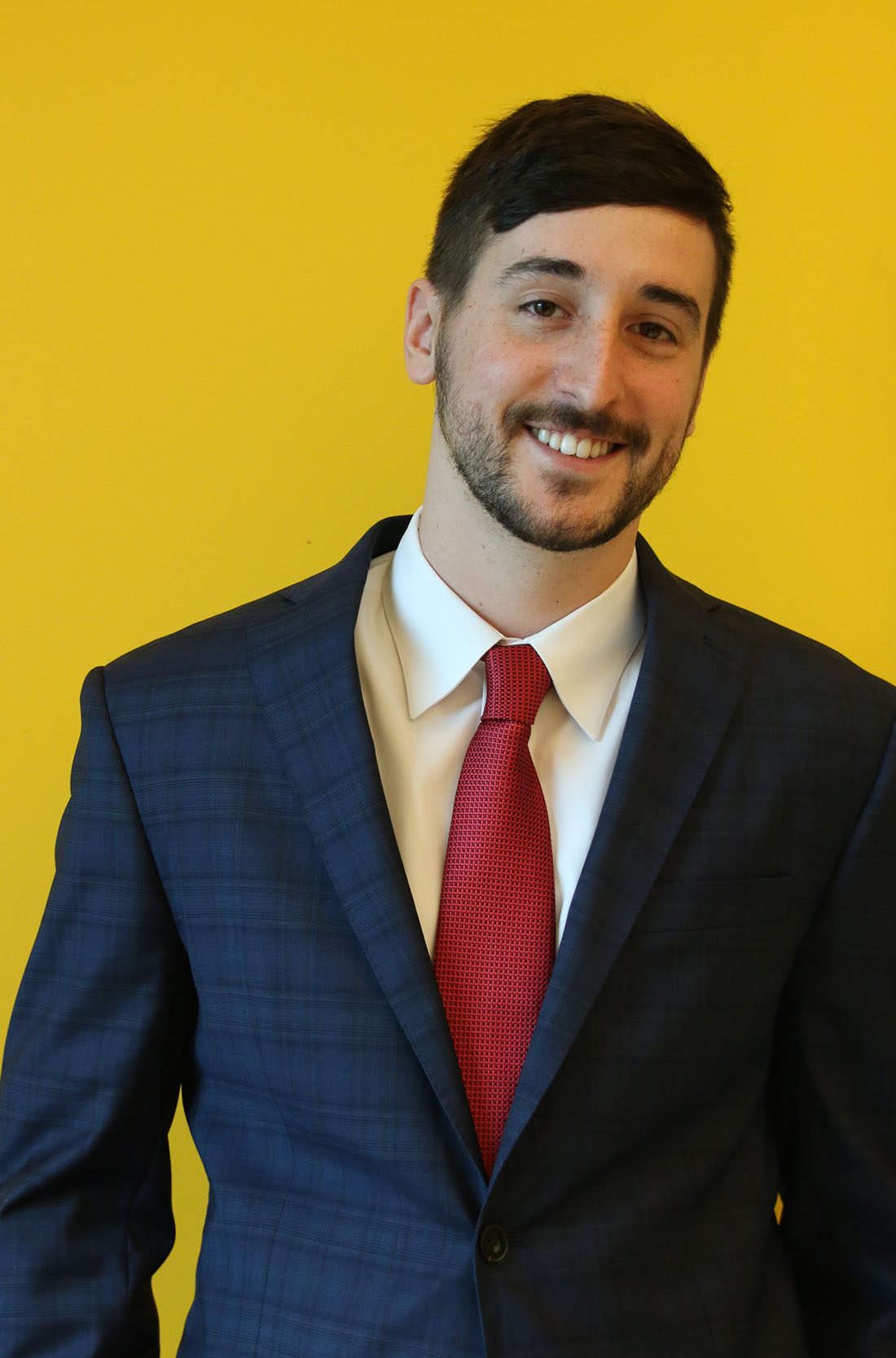 Jordan Mazurek is a young, white man with black side-swept hair and facial hair. He stands, smiling, against a yellow background wearing a navy suit jacket, white button up, and red tie.