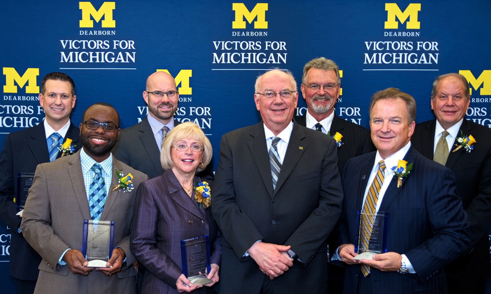7 Alumni Difference Makers pose with the Chancellor in front of UM-Dearborn Victors for Michigan banner.