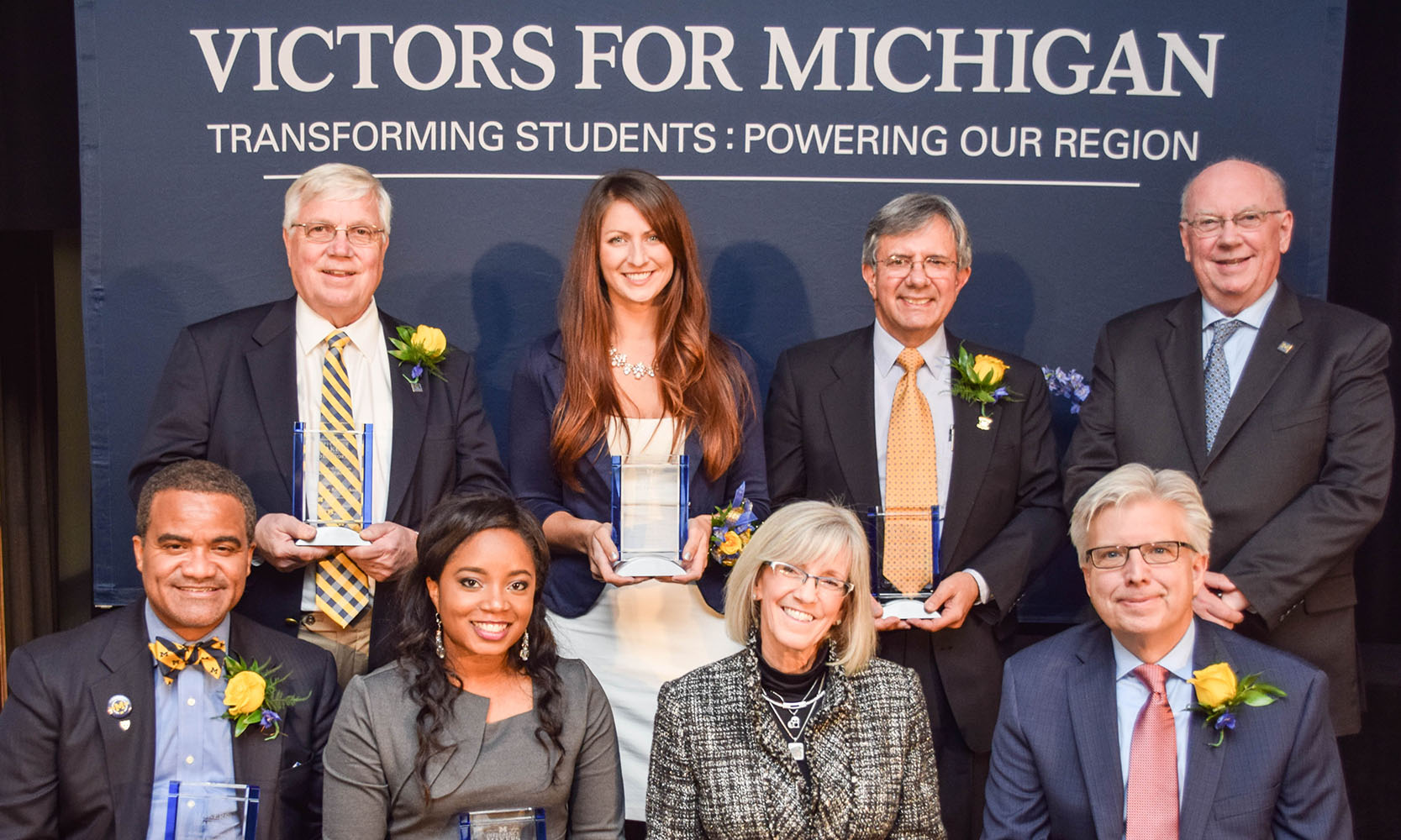 7 Alumni Difference Makers pose with the Chancellor in front of a Victors For Michigan, Transforming Students: Powering Our Region banner.