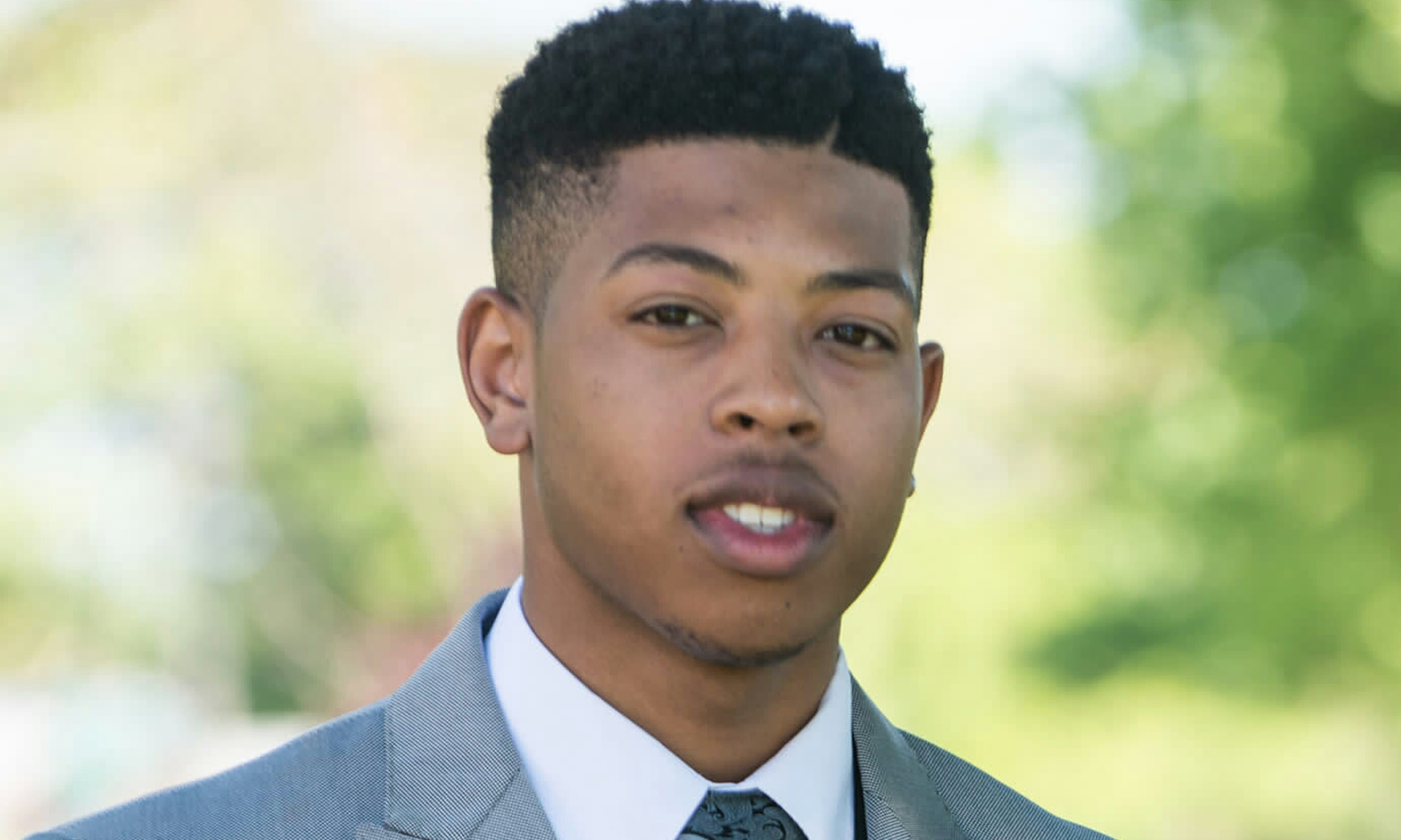 Jewell Jones is a young Black man with short, black, & faded hair and light facial hair. He is wearing a light blue suit jacket with a white button up and black tie.
