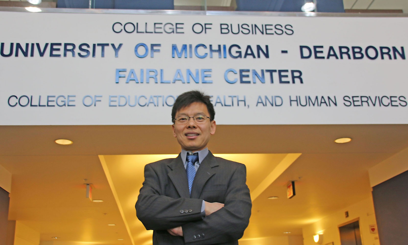 Jun He standing in front of wall that reads: College of Business, University of Michigan-Dearborn, Fairlane Center, College of Education, Health, and Human Services