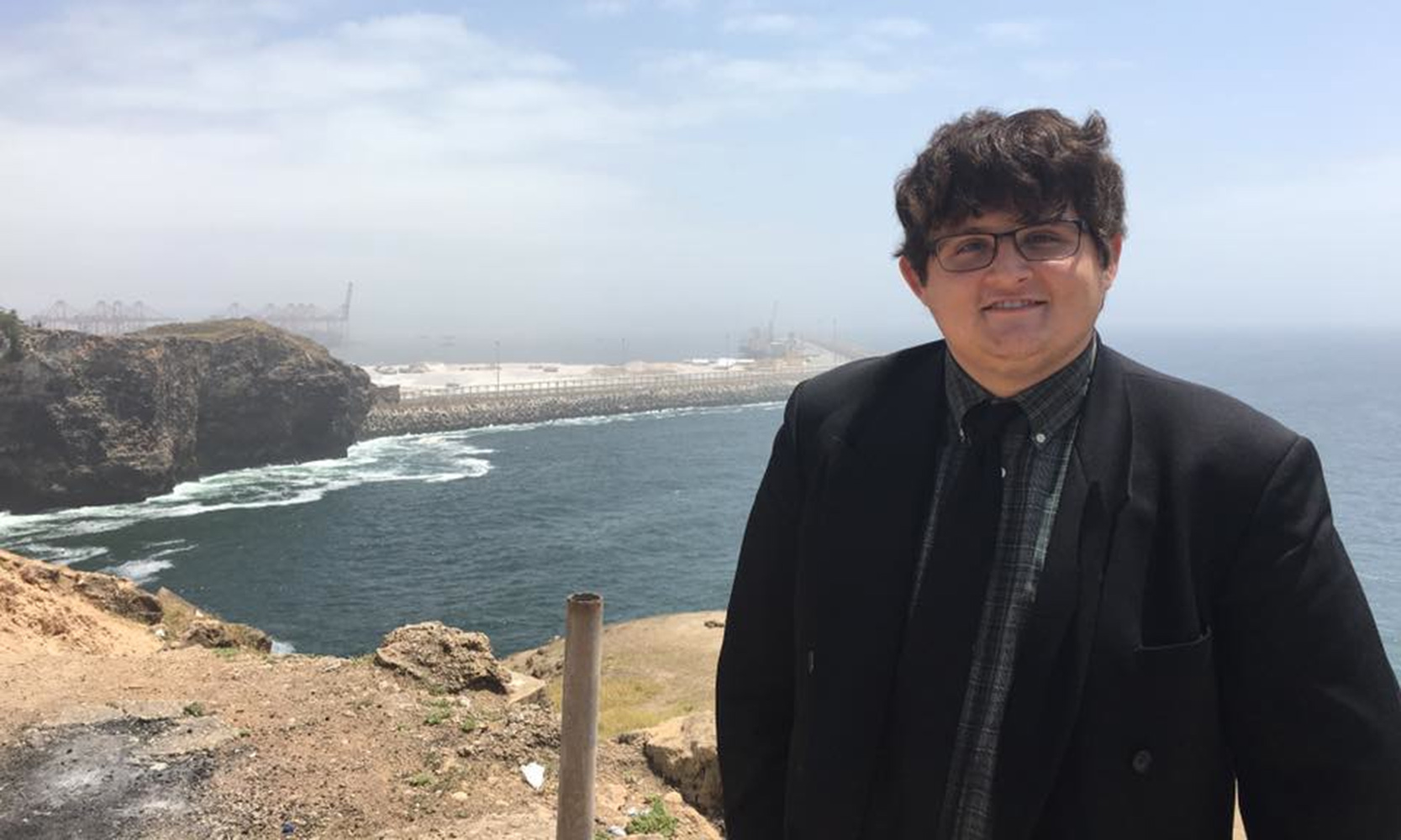 Christian Ledford is a young white man with brown hair hanging over his forehead and rectangular glasses. He stands in front of a sea in Oman wearing a dark green plaid button up with a black tie and black suit jacket.