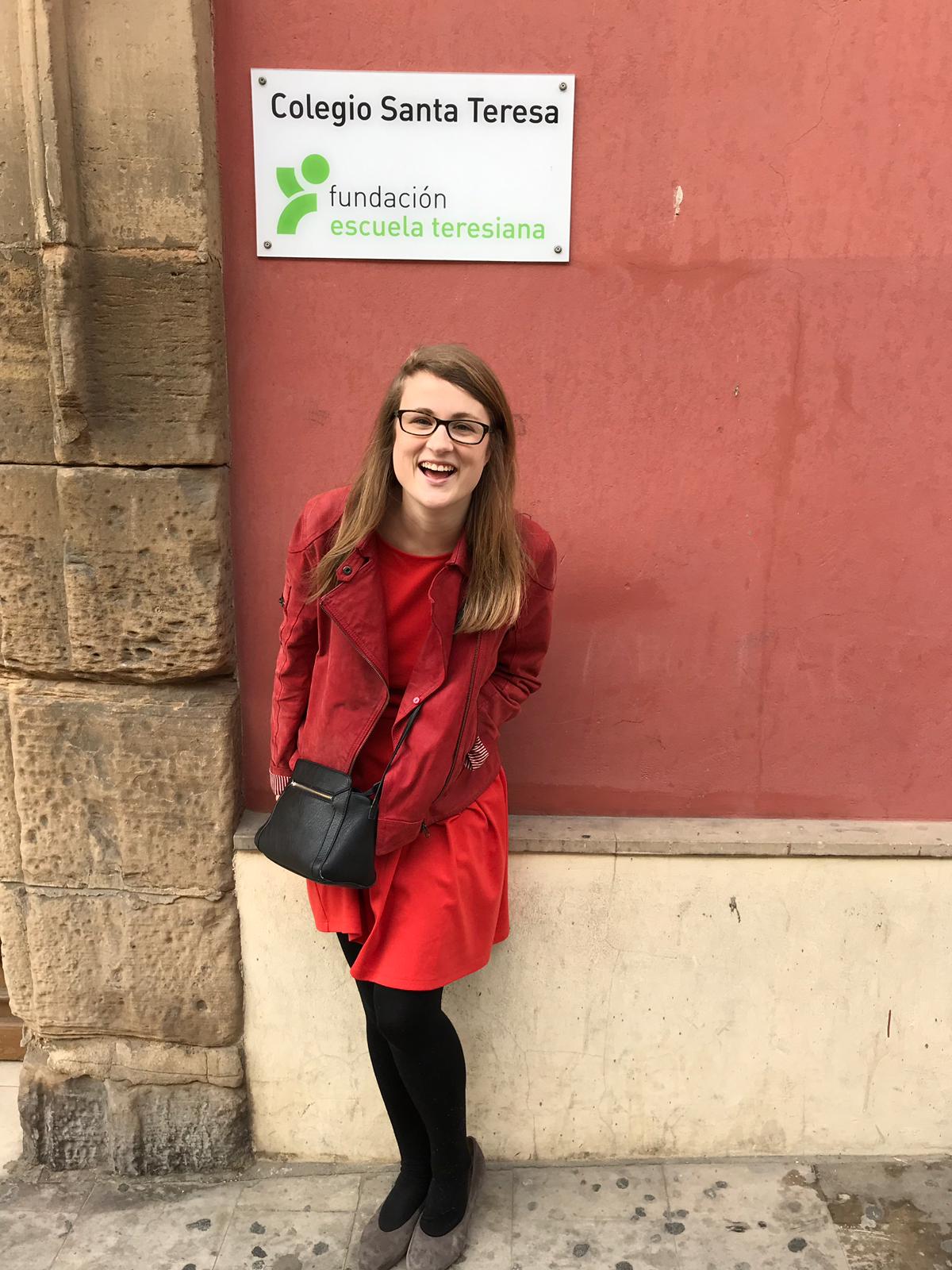 Erika MacLaren, smiling, in a read coat, standing outside the school where she taught English in Spain.