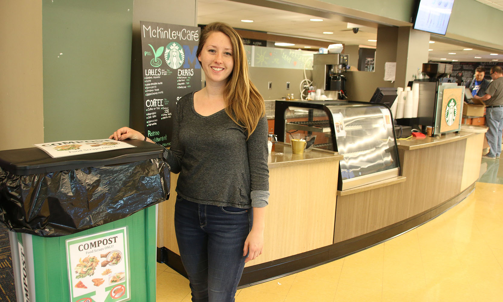 Sarah Bellaire is a young, white woman with straight, dark blonde hair that lays past her shoulders. She is wearing a dark, heather gray long-sleeve shirt and a pair of dark denim skinny jeans. She stands next to a green compost bin inside of the McKinley Cafe.