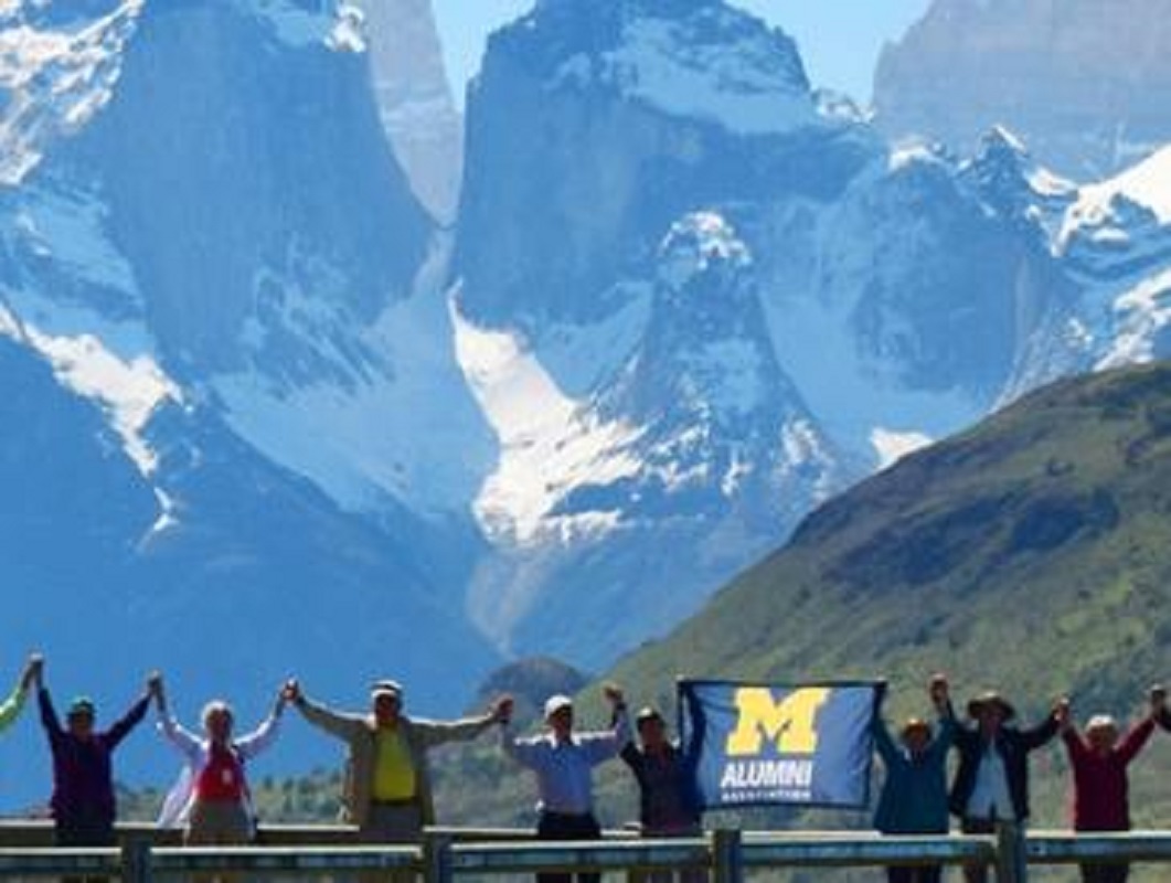 Alumni hold hands high and a UM-Dearborn Alumni flag stand at the base of snow mountain tops behind them.