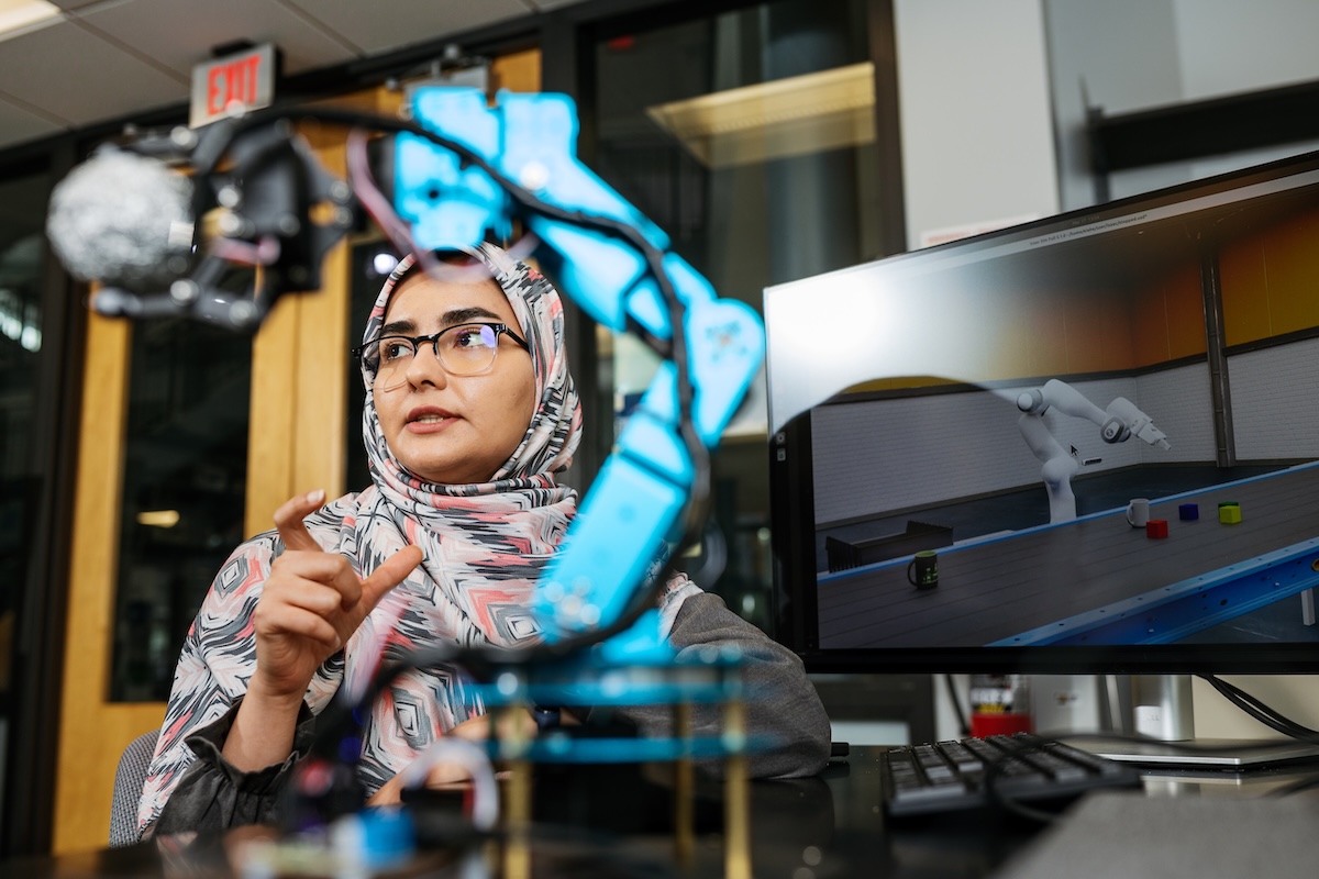A woman, who is wearing glasses and a hajab, stands behind a robotic arm in the lab.