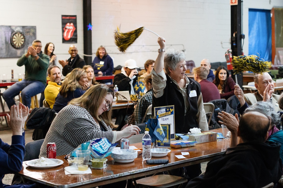 A group of people cheer for each other during a trivia game night. A woman shakes a maize-and-blue pom pom  over her head. 