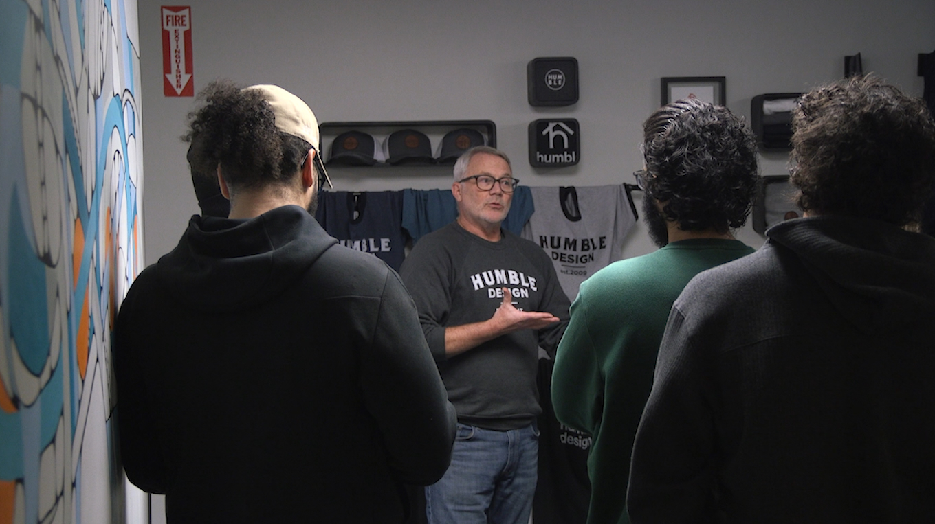 A man wearing a Humble Design shirt speaks to a group of students