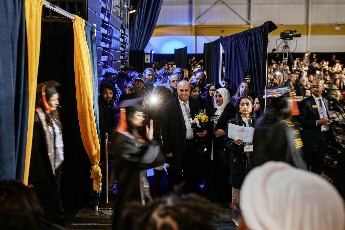 A large audience cheers and smiles when students wearing caps and gowns walk into the room.