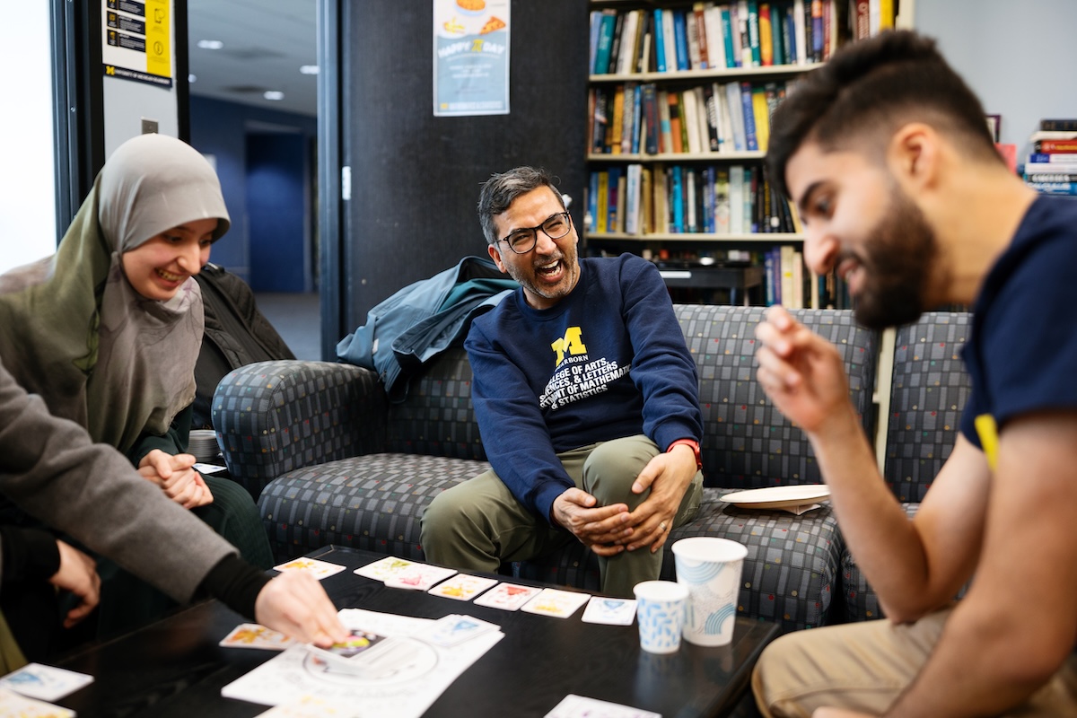 A professor, who is sitting on a couch and playing a card game, laughs with his students.