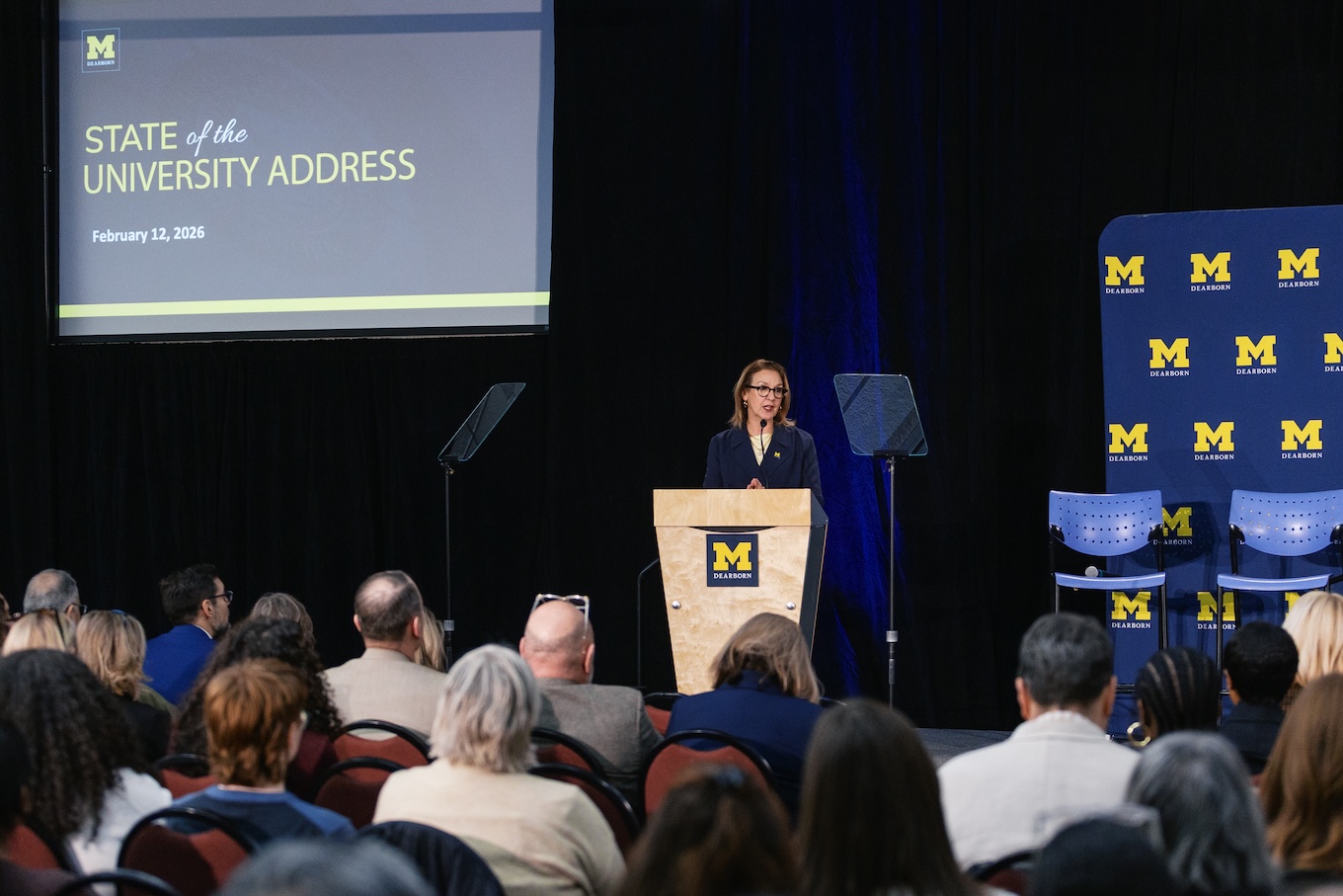 A woman wearing a navy blue suit stands behind a podium to deliver a speech.