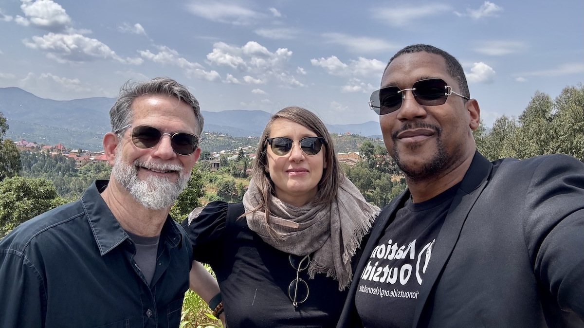 Three people, two males and a female, are smiling in an outdoor photo taken in Rwanda. They are on the top of a mountain with a city and trees in the background.