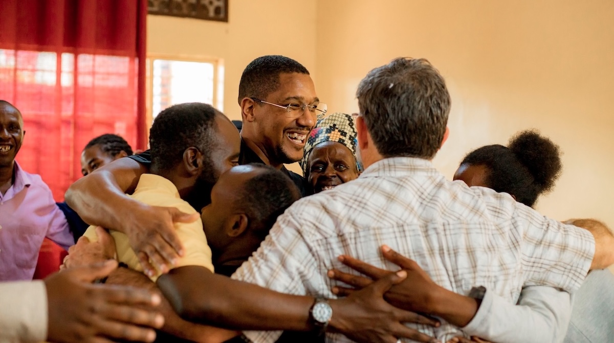 A smiling man with glasses is hugged by a group of happy people. They are celebrating forgiveness and community building.