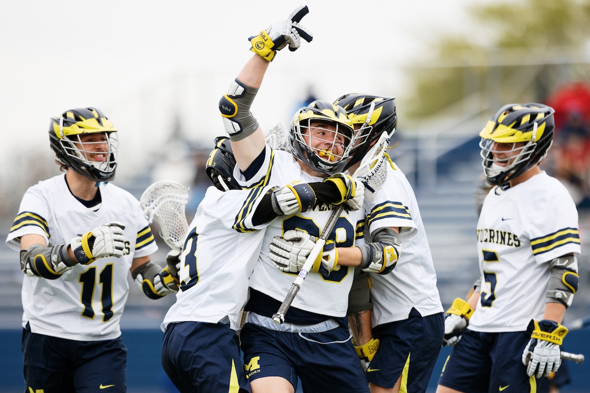 UM-Dearborn men's lacrosse team members hug one another and cheer after scoring a goal. They are wearing maize and blue helmets and white jerseys with maize and blue accents.