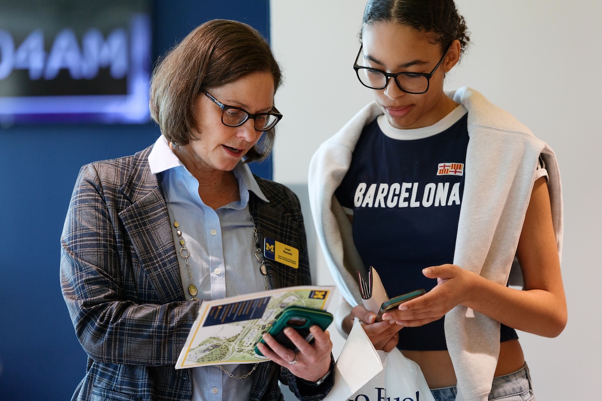 Two women - one professor and one student - talk about the majors UM-Dearborn offers at a campus event. The professor has on a plaid jacket and a light blue shirt. The student is wearing a navy blue Barcelona football shirt with a sweater around her shoulders.