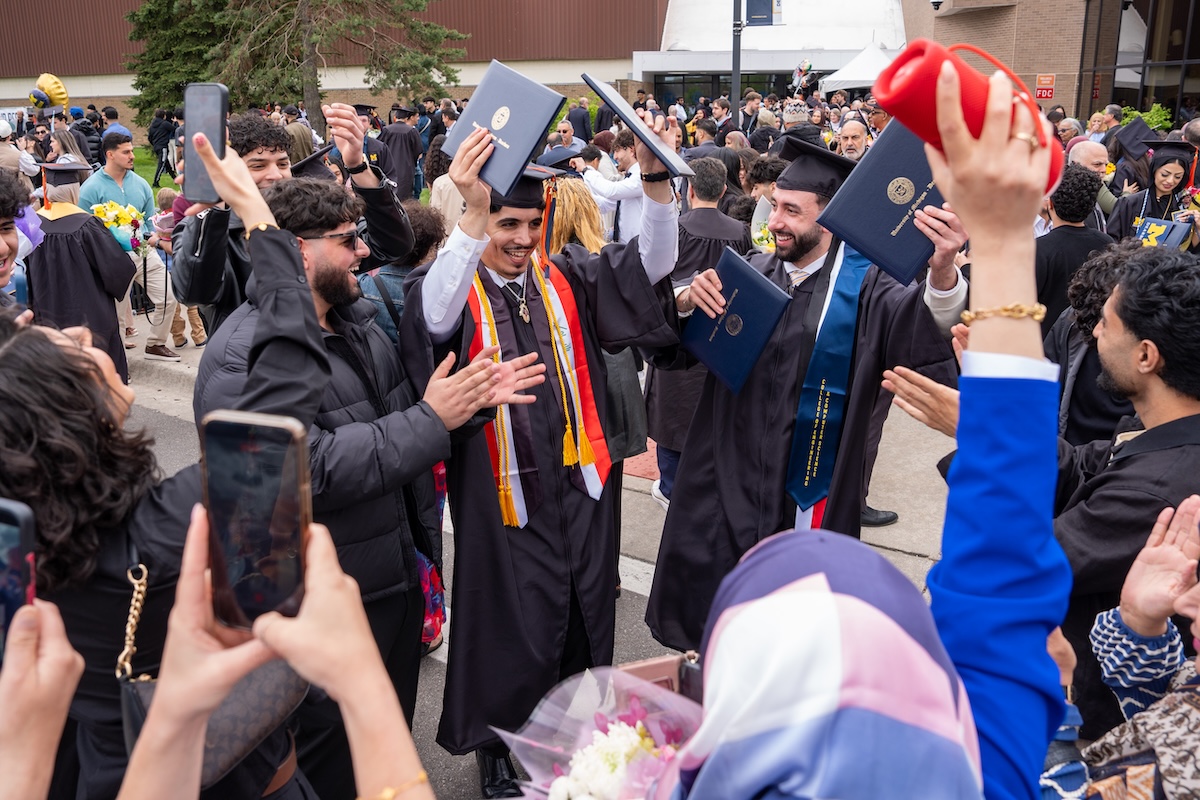 A group of men wearing caps and gowns celebrate their graduation by holding their blue diploma covers into the air. 