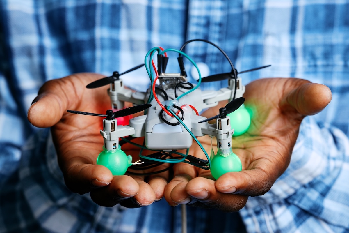 Close-up view of a person wearing a blue plaid shirt holding a small, custom-built drone with open palms. The drone has four propellers, exposed red, blue, and green wiring, and bright green rounded covers on its landing gear.