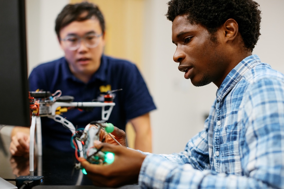 A young man in a blue plaid shirt inspects a small, illuminated drone in a laboratory while a second person in a navy shirt watches in the background.