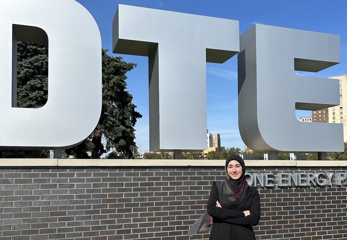 A woman wearing a black headscarf and blazer standing with crossed arms in front of large, 3D grey letters spelling "DTE" mounted on a brick wall.