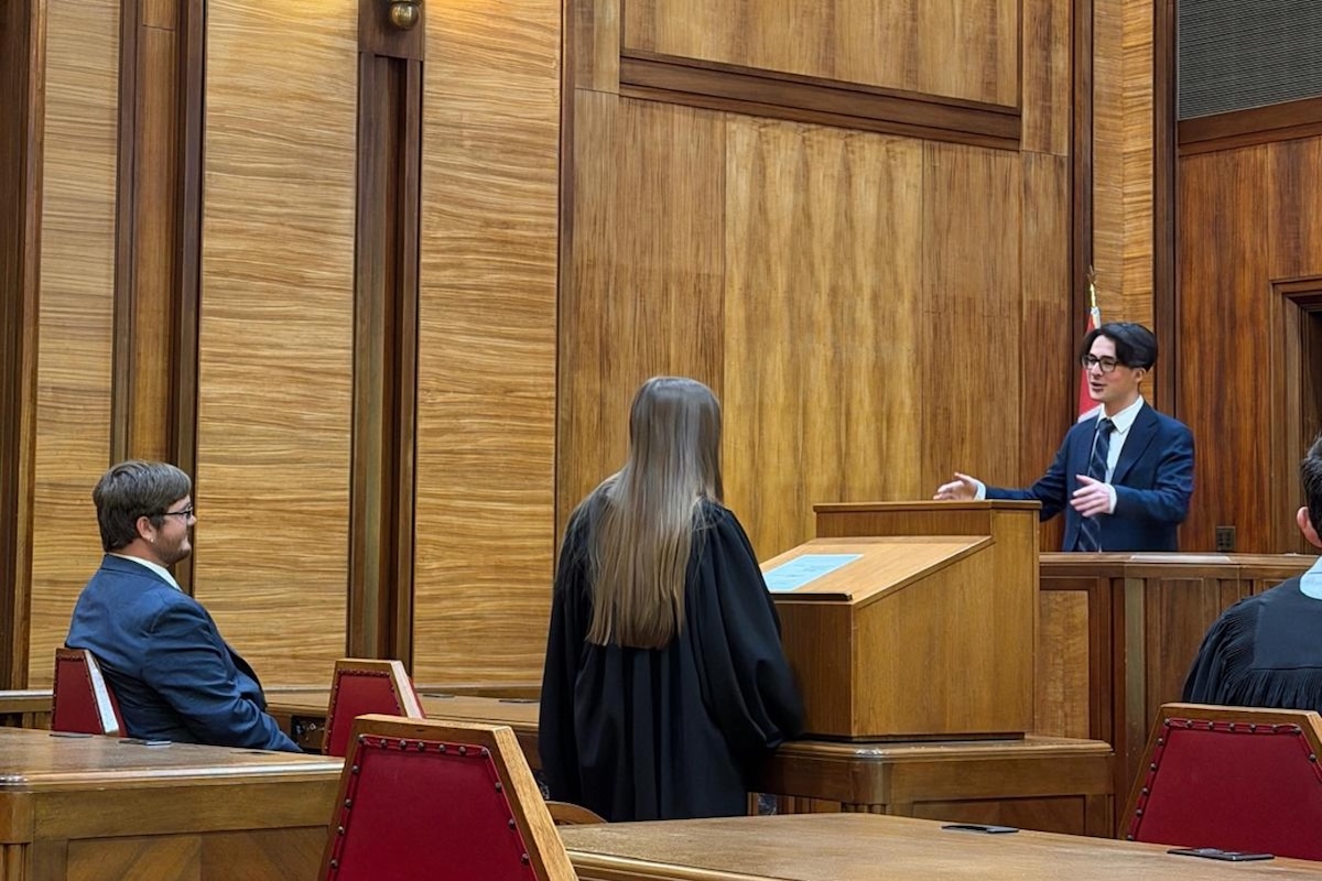 A young man in a dark suit speaks at a witness stand or bench in a wood-paneled courtroom while a person in black judicial robes stands at a podium facing him.