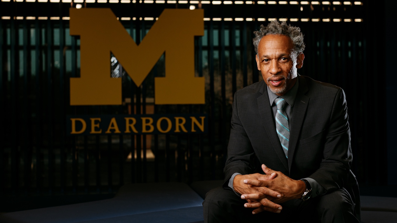 A man seated and staring at the camera in front of the UM-Dearborn Block M