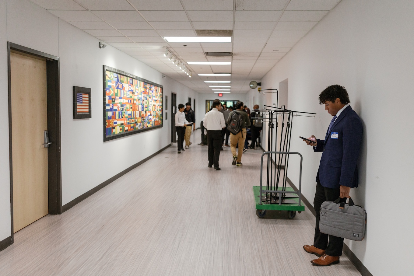A male student looks at his phone while standing in a hallway while a group of students walk past him.