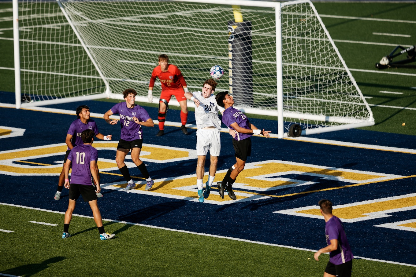 A group of male soccer players gather around the white goal net, and a UM-Dearborn uses his head to bump the soccer ball.