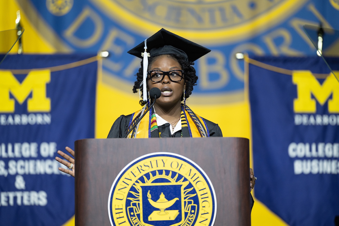 A woman wearing a cap and gown speaks at a podium with a maize and blue sign in the background