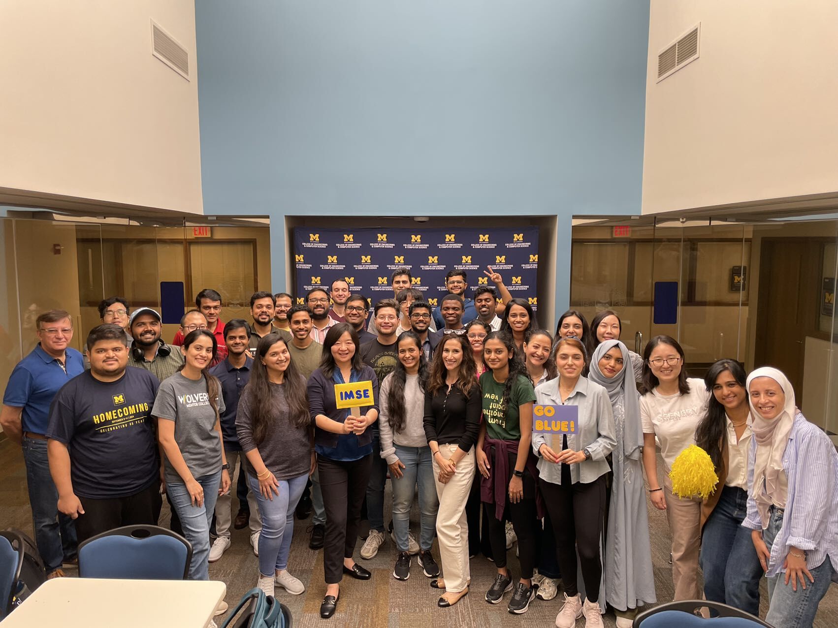 Large group of students pose together indoors with University of Michigan signs, including “IMSE” and “Go Blue,” and many are wearing maize and blue apparel.