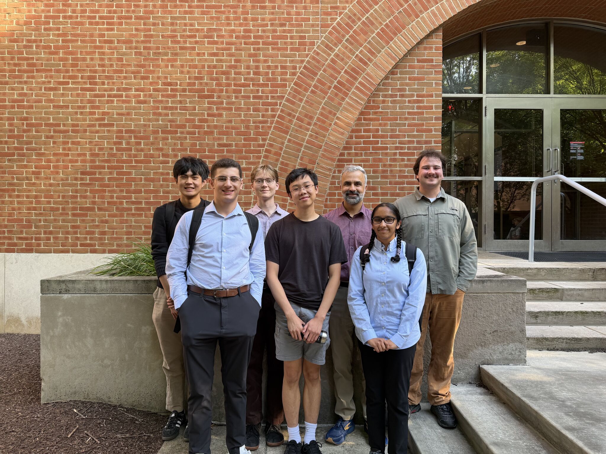 A group of undergraduate REU students and faculty mentor Professor Yunus Zeytuncu pose for a photo outside the Natural Science Building at the University of Michigan-Dearborn. The students smile together in front of the building's brick archway, representing a collaborative summer mathematics research experience.