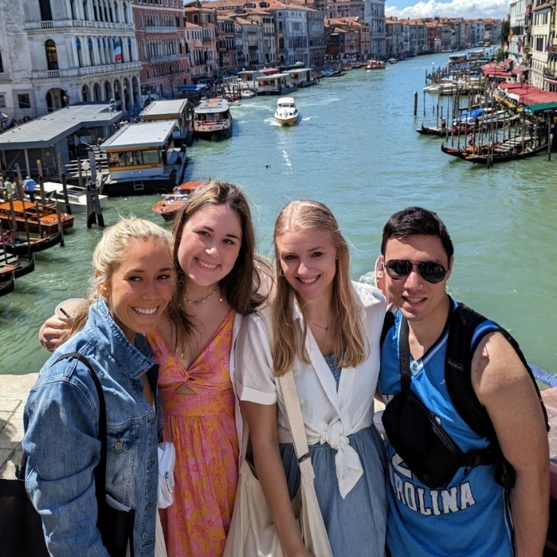 Group of four smiling people standing on a bridge over a canal in Venice with boats and historic buildings behind them.