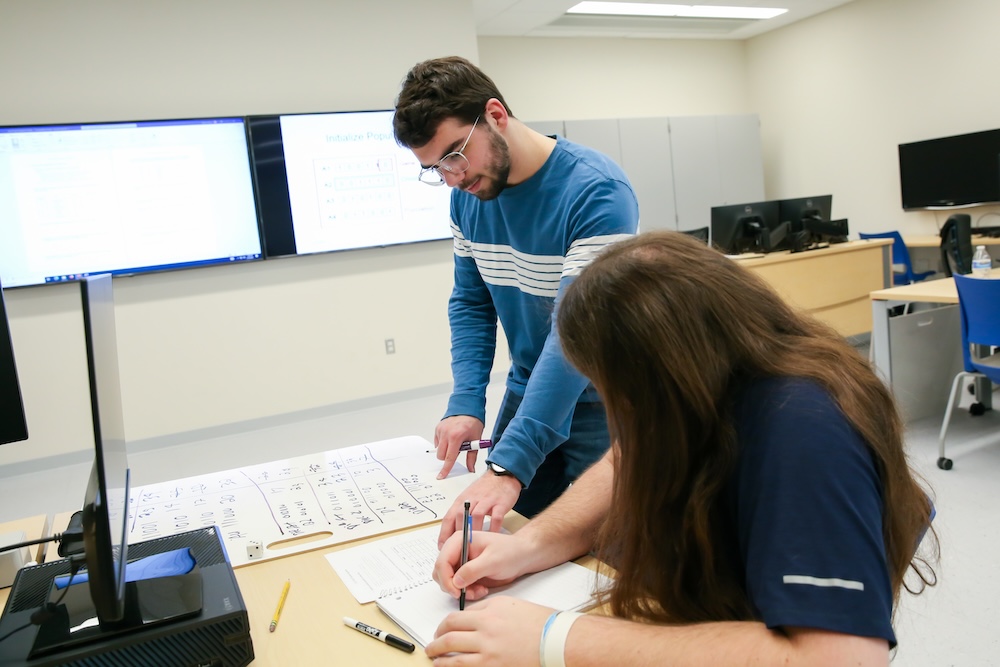 Two individuals collaborate at a table with large sheets of paper, with one person writing and the other observing. In the background, a computer screen displays a flowchart.
