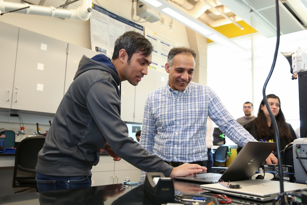 Two individuals are collaborating over a laptop in a laboratory setting, surrounded by scientific equipment.