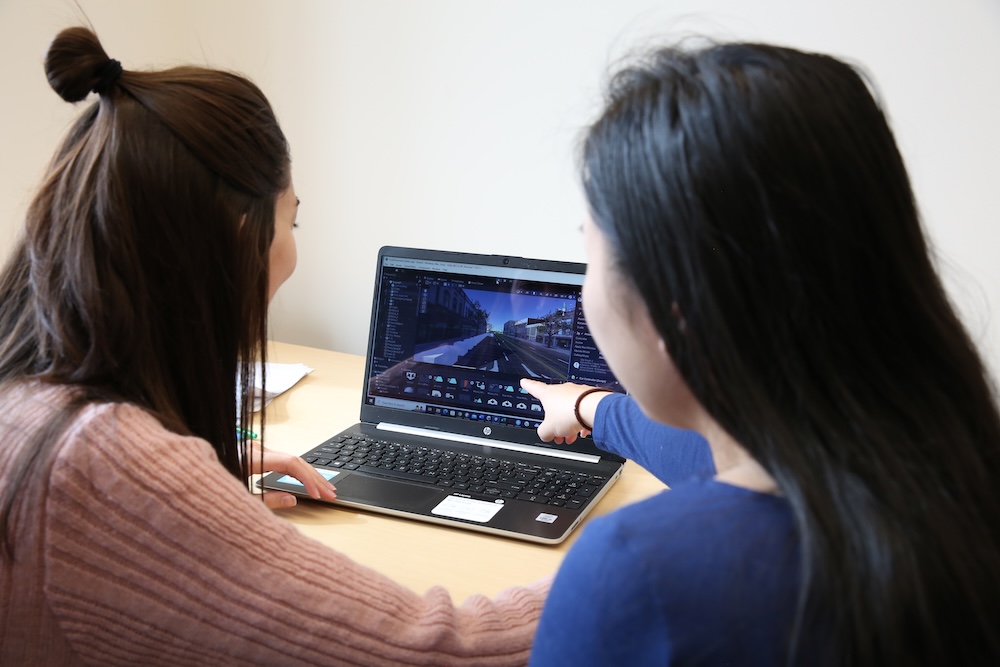 Two people are sitting at a desk and viewing a laptop screen displaying a video game scene.