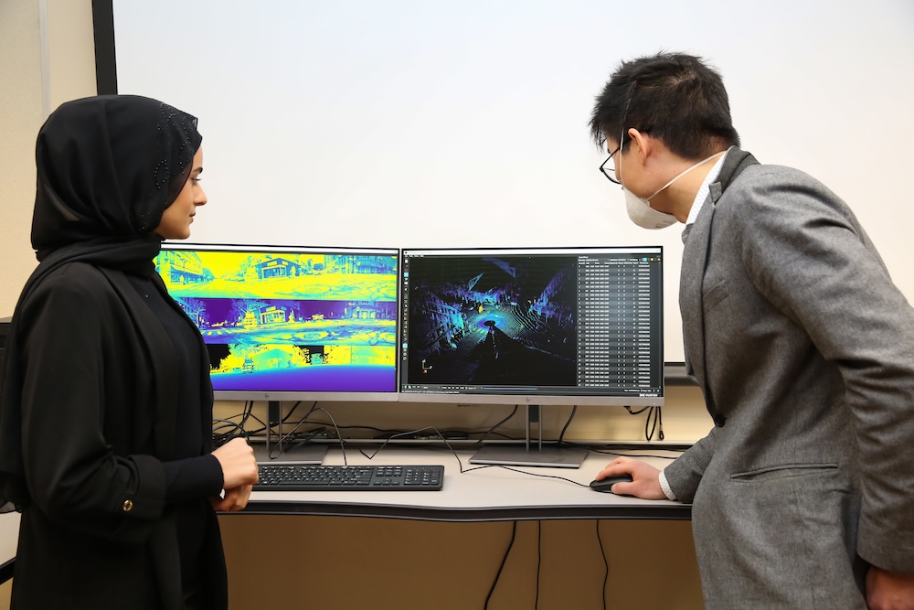 Two professionals, one wearing a hijab, stand beside a desk examining thermal images on computer monitors. They are engaged in a discussion in an office environment.
