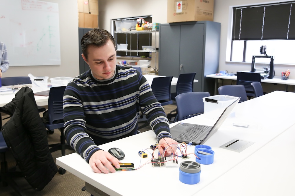 A person working on an electronic circuit board at a table with a laptop and electronic components in a classroom environment.